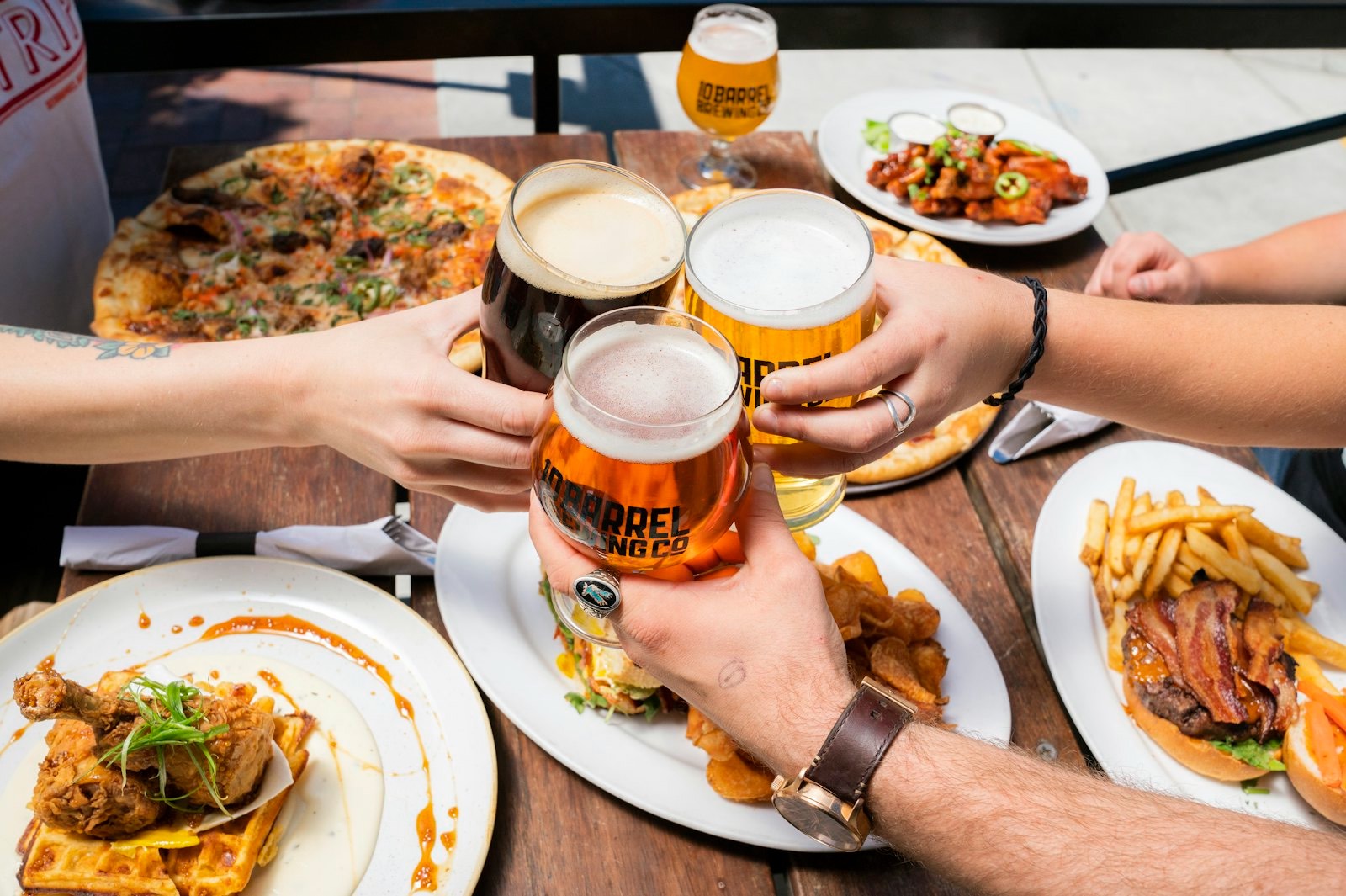Friends clinking craft-beer glasses over a table of pizza, a burger and wings